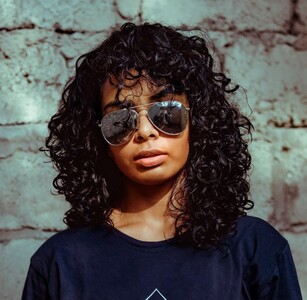 A imagem mostra uma moça negra, de óculos de sol, com um corte de cabelo bem repicado na altura dos ombros.