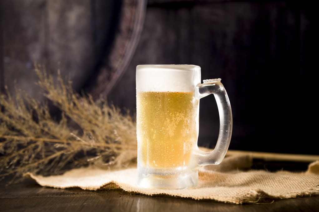 A imagem mostra uma caneca de cerveja gelada sobre uma mesa de madeira, com alguns ramos de trigo ao fundo.