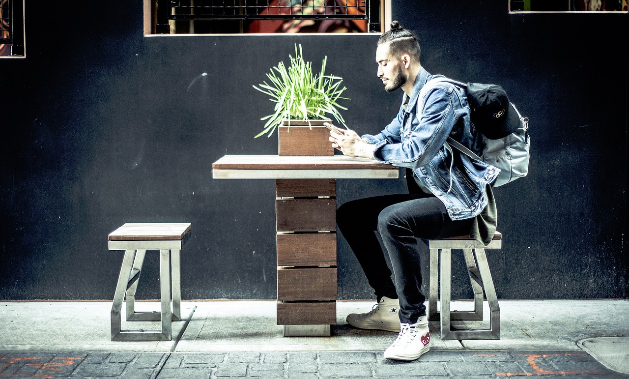 A imagem apresenta um homem jovem sentado em um banco frente a uma mesa, mexendo no celular.