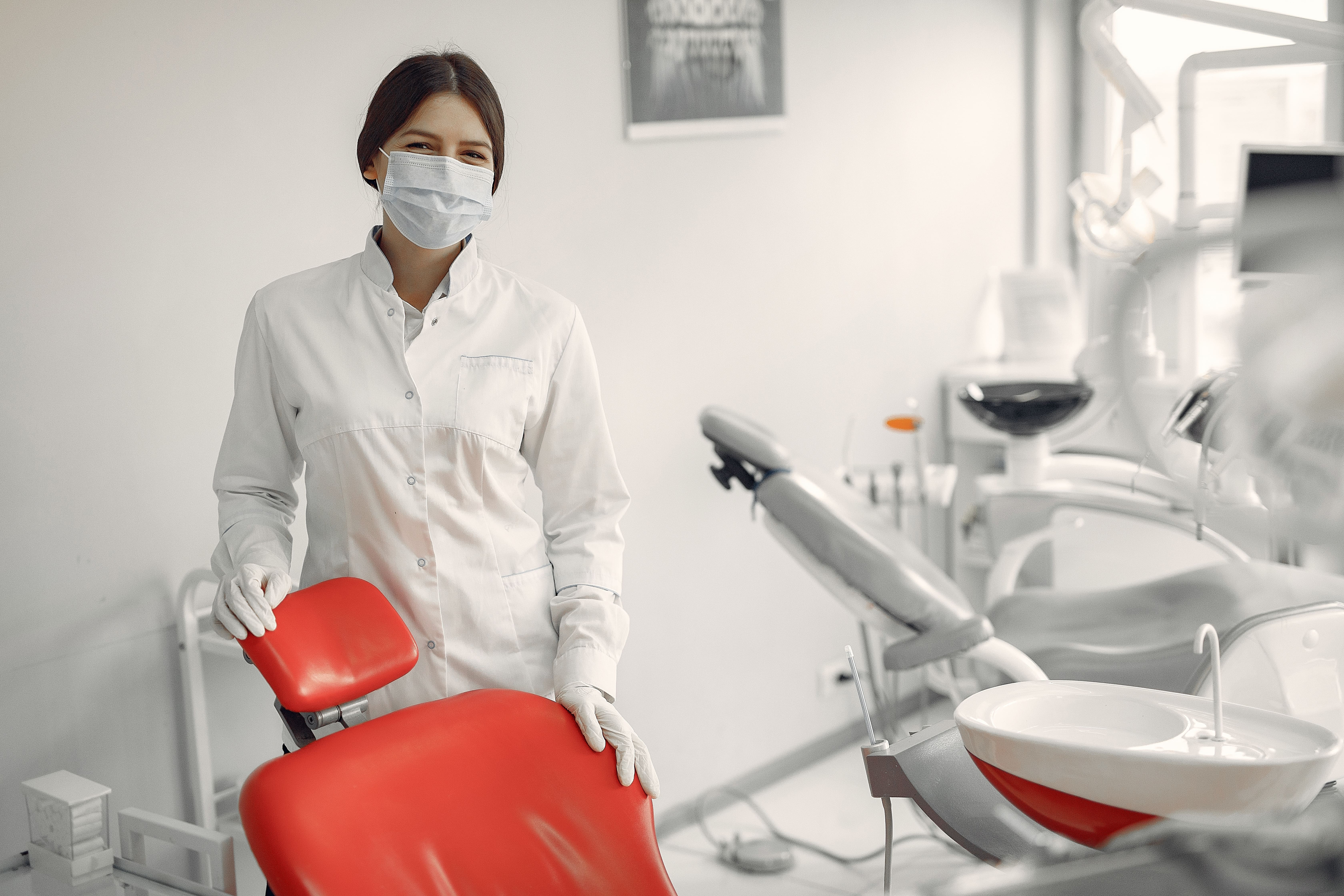 Mulher dentista está atrás de uma cadeira de consultório, olhando para a câmera e sorrindo. 