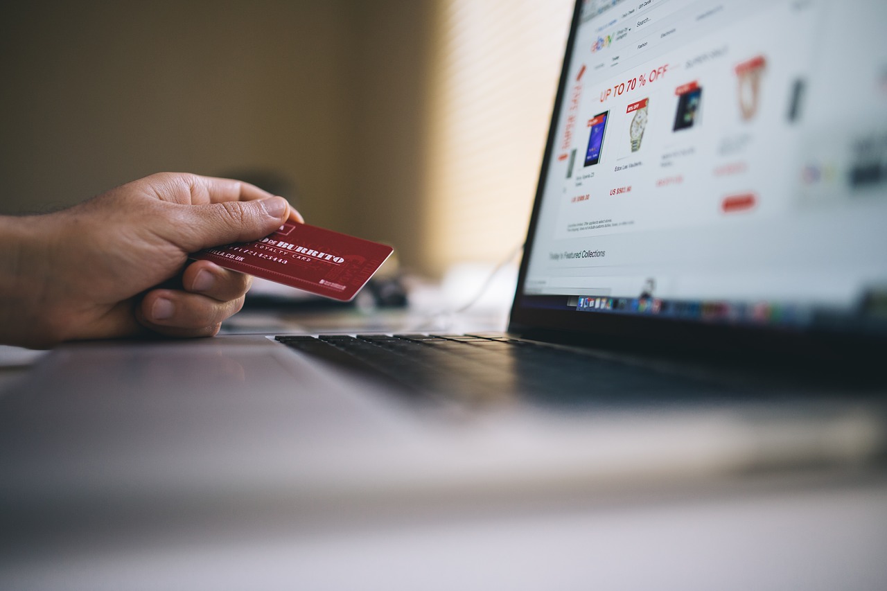 A imagem mostra uma pessoa em frente a um computador, cuja tela mostra um marketplace com vários produtos. Na mão dessa pessoa é possível notar um cartão de crédito vermelho.