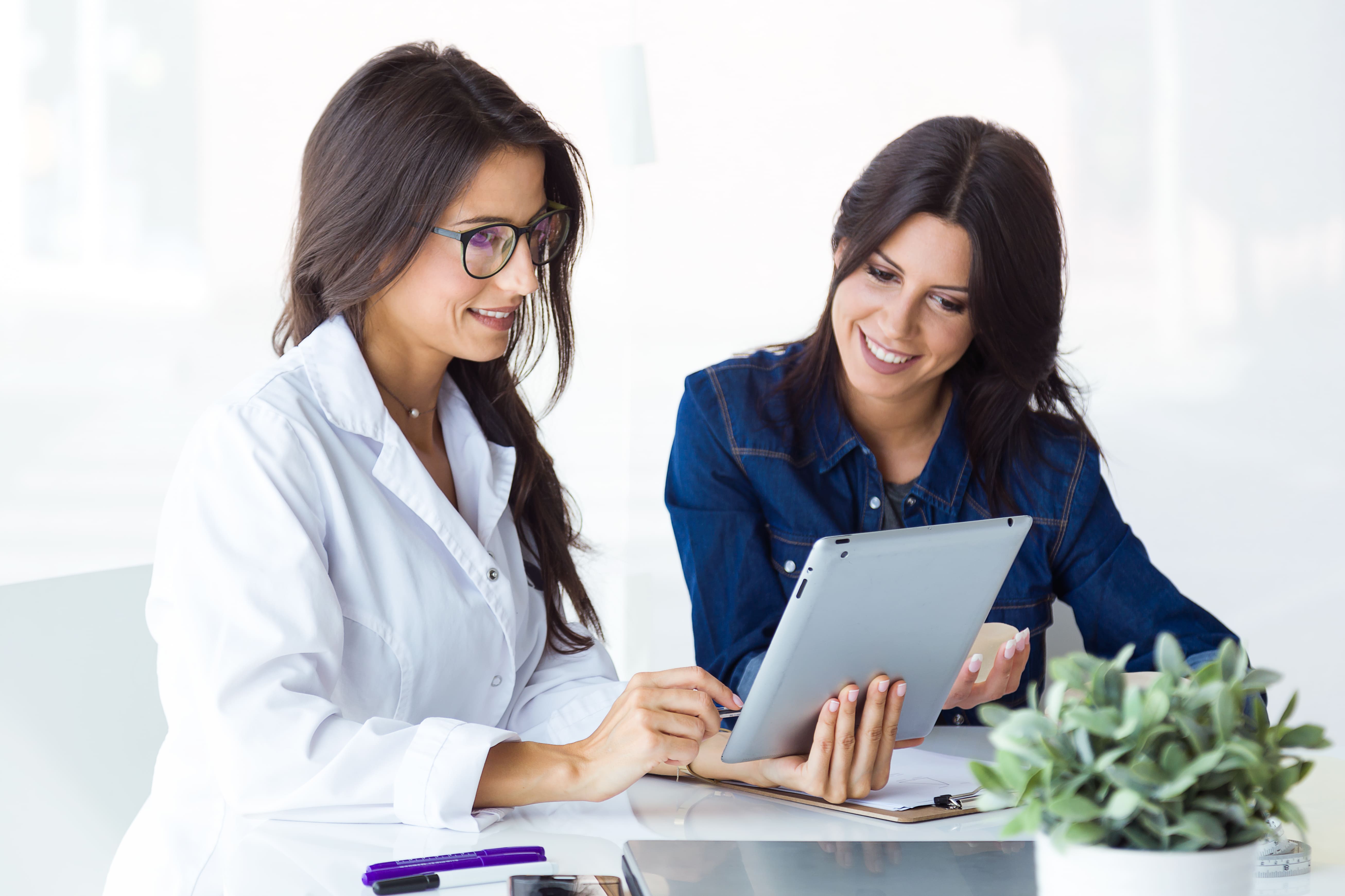 Há duas mulheres, uma é médica e outra é sua paciente. Ambas estão olhando para o tablet que a médica segura.