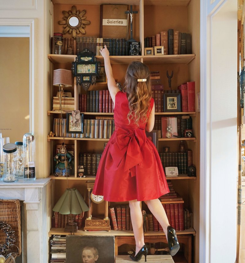 A imagem mostra uma mulher de vestido vermelho e cabelos castanhos organizando uma estante de livros. É possível observar vários objetos de enfeite em volta. 