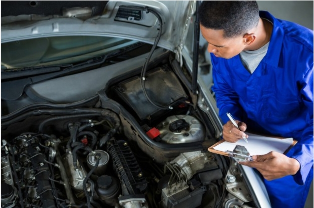 A imagem mostra um profissional realizando a revisão no motor de um carro. Ele está com um macacão azul e segura uma prancheta de madeira, na qual faz anotações.