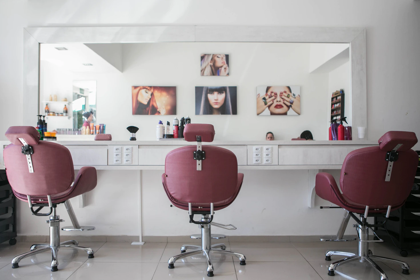  A imagem mostra um salão de beleza com paredes brancas com fotos de modelos penduradas. Há três cadeiras de clientes e um espelho grande.