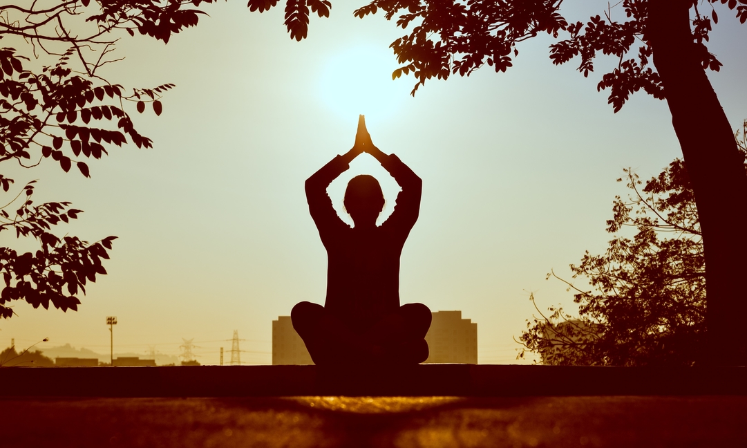 A imagem mostra uma mulher de costas realizando uma asana (postura) de yoga. A mesma se encontra sentada, com os joelhos cruzados na posição de lótus e com as mãos unidas acima da cabeça.